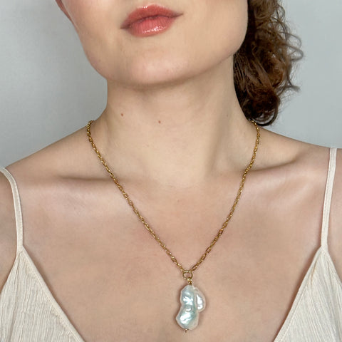 A close-up of a woman wearing a delicate gold chain necklace with a large, irregular white baroque pearl pendant, paired with a light beige camisole top against a neutral gray background.